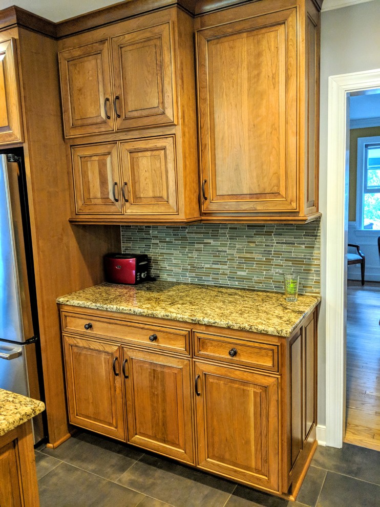 Inspiration for a mid-sized transitional u-shaped porcelain tile and black floor kitchen remodel with a farmhouse sink, raised-panel cabinets, light wood cabinets, granite countertops, green backsplash, glass sheet backsplash, stainless steel appliances, an island and brown countertops