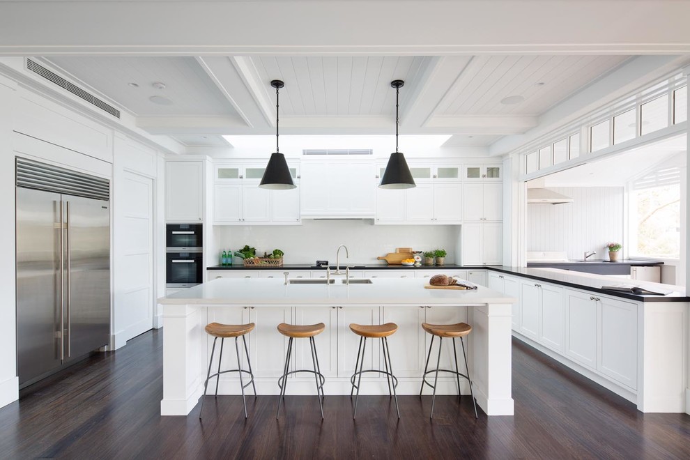 Photo of an expansive beach style l-shaped open plan kitchen in Sydney with an undermount sink, shaker cabinets, white cabinets, quartz benchtops, white splashback, subway tile splashback, stainless steel appliances, dark hardwood floors, with island, brown floor and white benchtop.