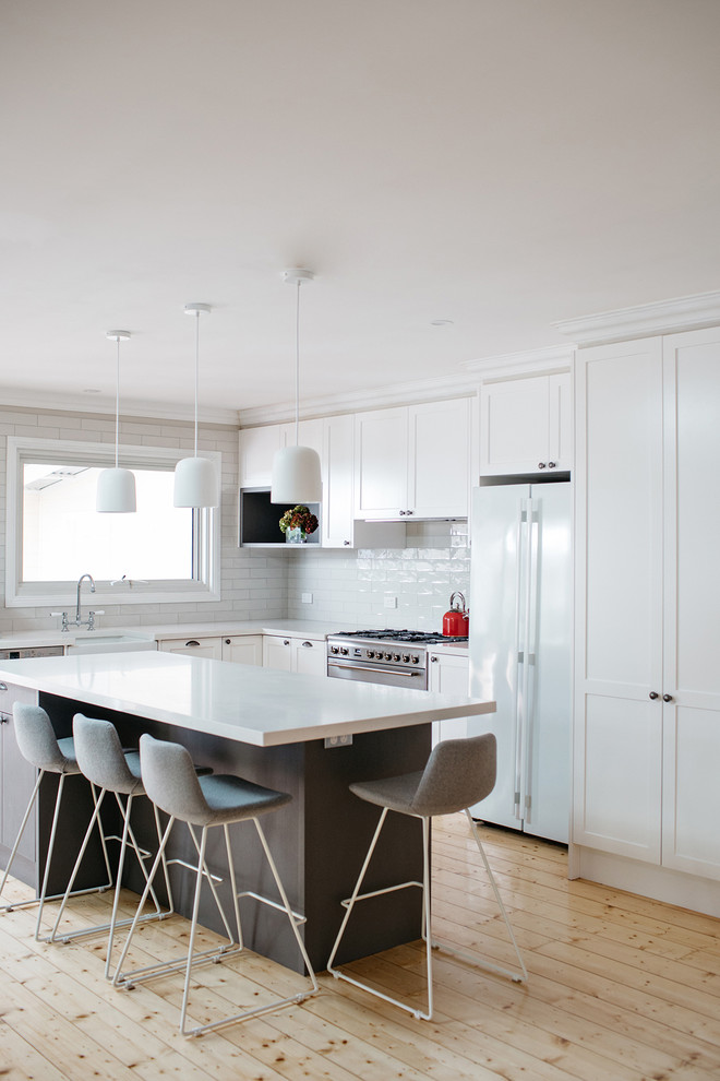 Photo of a large transitional l-shaped open plan kitchen in Melbourne with a farmhouse sink, shaker cabinets, white cabinets, quartz benchtops, white splashback, subway tile splashback, stainless steel appliances, light hardwood floors and with island.