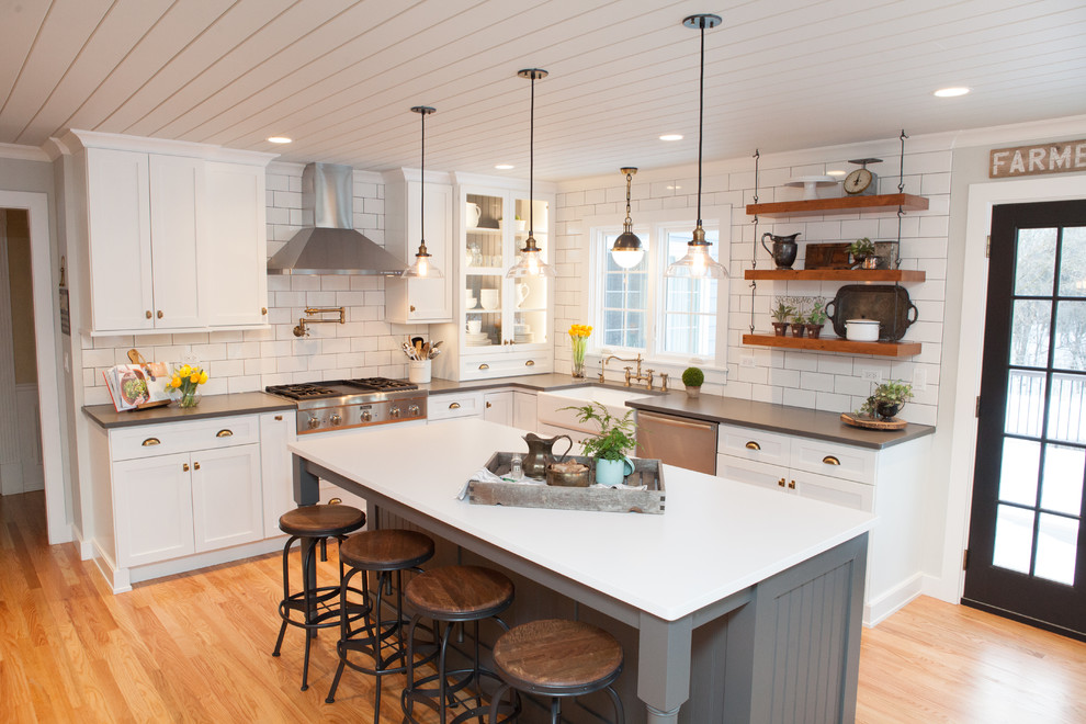 Design ideas for a country l-shaped kitchen in Chicago with a farmhouse sink, shaker cabinets, white cabinets, white splashback, subway tile splashback, stainless steel appliances, light hardwood floors and with island.