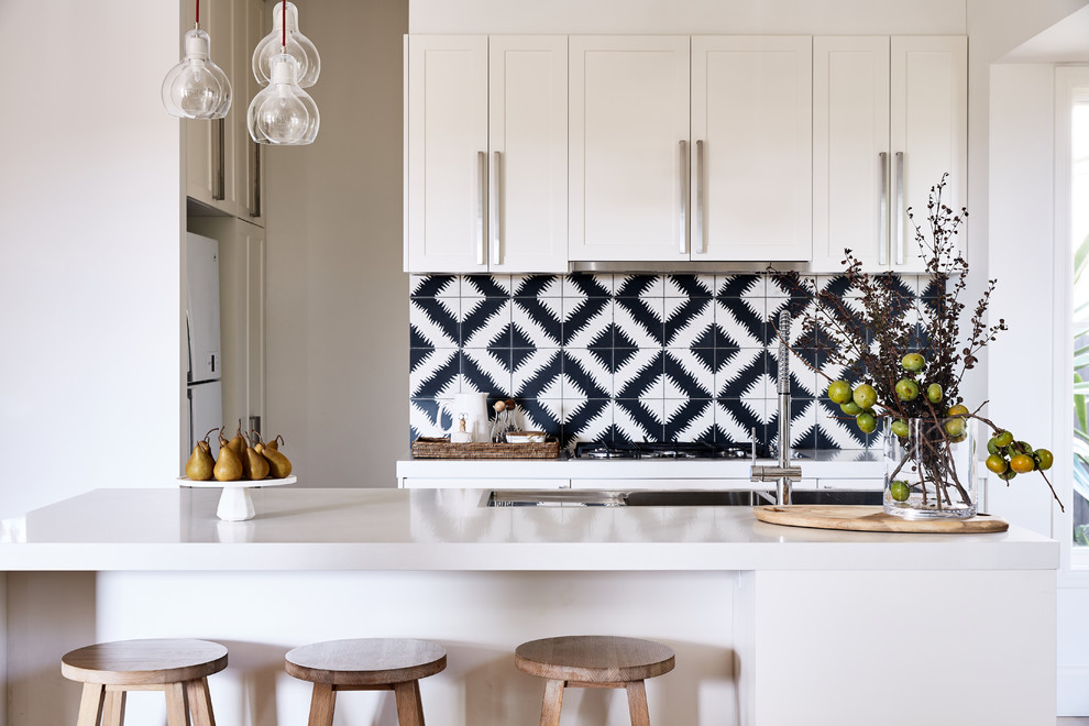 This is an example of a transitional l-shaped kitchen in Melbourne with a double-bowl sink, shaker cabinets, white cabinets, quartz benchtops, cement tile splashback, stainless steel appliances, light hardwood floors, with island, white benchtop and black splashback.