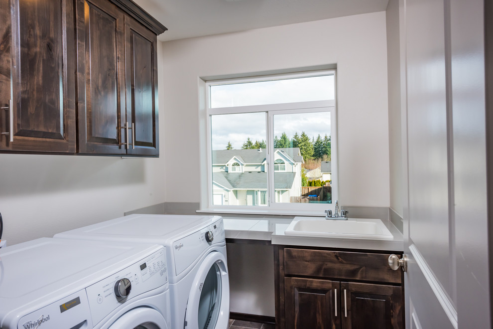 This is an example of a contemporary dedicated laundry room in Portland with a drop-in sink, raised-panel cabinets, dark wood cabinets, tile benchtops, white walls, vinyl floors and a side-by-side washer and dryer.