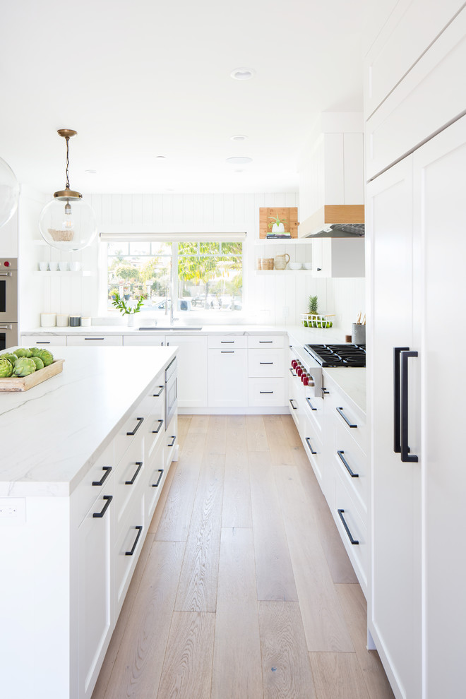 This is an example of a beach style l-shaped eat-in kitchen in Orange County with an undermount sink, shaker cabinets, white cabinets, quartzite benchtops, white splashback, timber splashback, panelled appliances, light hardwood floors, with island and white benchtop.