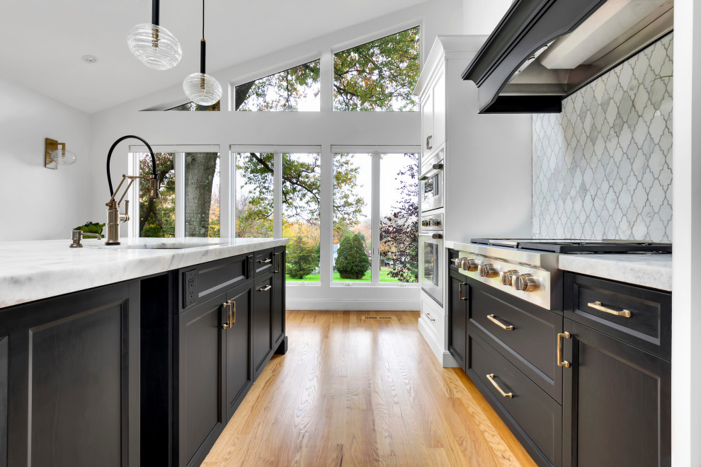 This is an example of a large transitional l-shaped eat-in kitchen in New York with an undermount sink, shaker cabinets, black cabinets, quartz benchtops, white splashback, marble splashback, panelled appliances, light hardwood floors, with island, beige floor, white benchtop and vaulted.