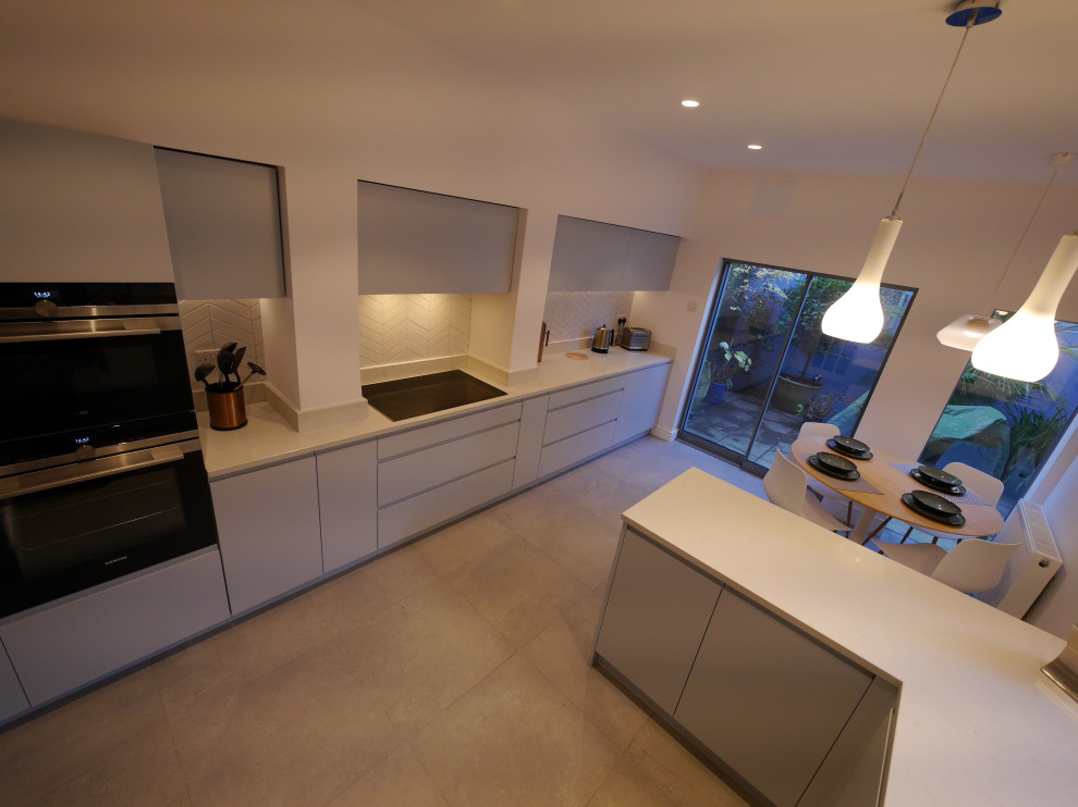 Photo of a mid-sized contemporary galley separate kitchen in London with an undermount sink, flat-panel cabinets, blue cabinets, quartzite benchtops, white splashback, ceramic splashback, black appliances, ceramic floors, a peninsula, grey floor and white benchtop.