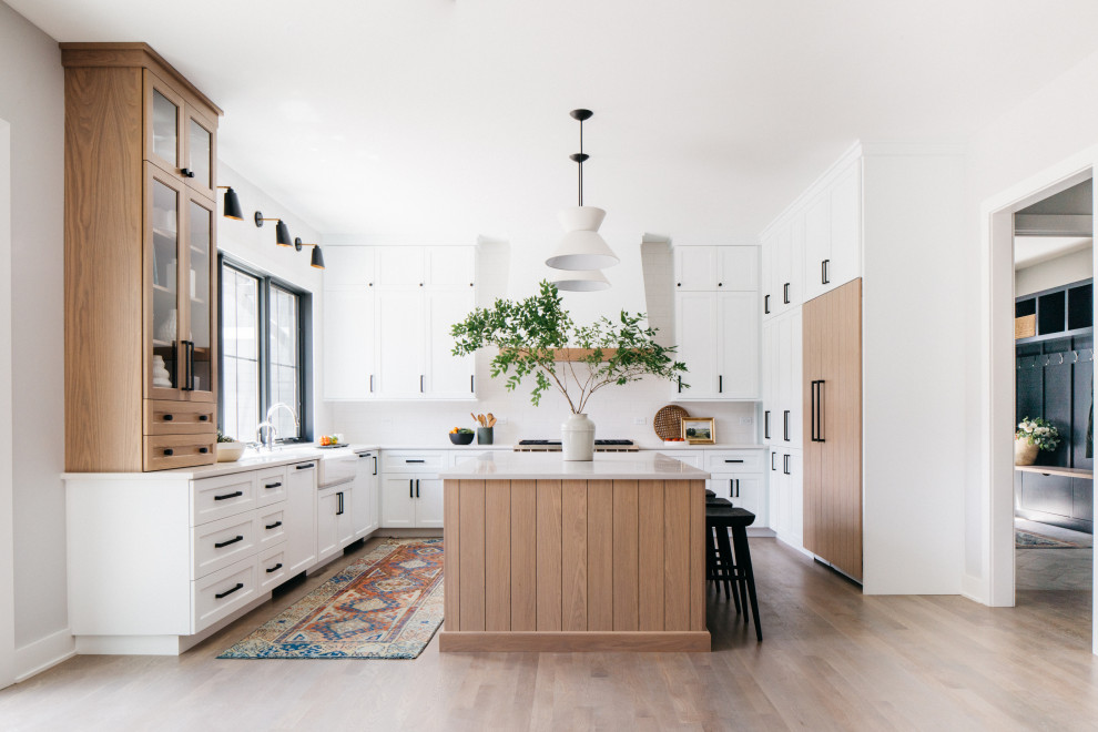Transitional kitchen in Chicago with a farmhouse sink, shaker cabinets, white cabinets, white splashback, panelled appliances, light hardwood floors, with island, brown floor and white benchtop.