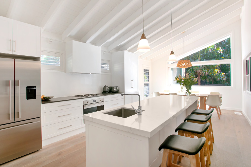 Beach style galley eat-in kitchen in Central Coast with a single-bowl sink, shaker cabinets, white cabinets, white splashback, glass sheet splashback, stainless steel appliances, light hardwood floors, with island, beige floor and white benchtop.