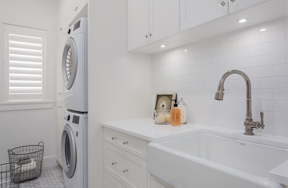 Inspiration for a mid-sized country single-wall dedicated laundry room in Vancouver with a farmhouse sink, shaker cabinets, white cabinets, quartz benchtops, white splashback, subway tile splashback, white walls, light hardwood floors, a stacked washer and dryer, grey floor, white benchtop and planked wall panelling.