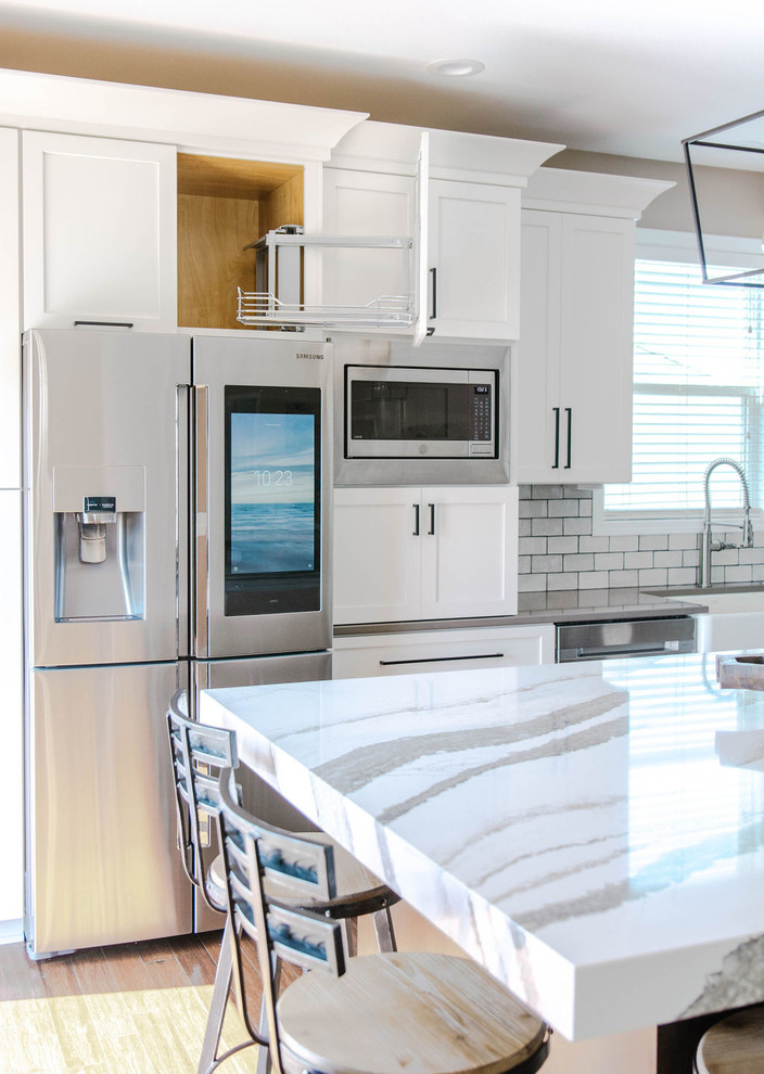 Example of a mid-sized transitional l-shaped medium tone wood floor and brown floor eat-in kitchen design in Omaha with a farmhouse sink, shaker cabinets, white cabinets, quartzite countertops, white backsplash, subway tile backsplash, stainless steel appliances, an island and white countertops