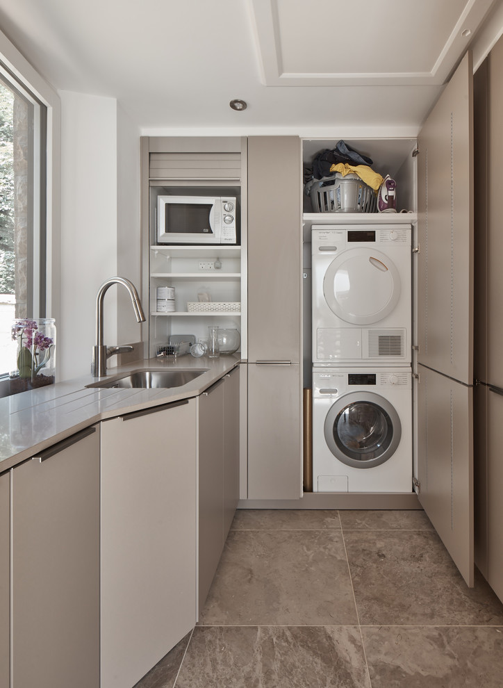Design ideas for a mid-sized contemporary single-wall laundry room in Other with flat-panel cabinets, solid surface benchtops, brown splashback, mirror splashback, limestone floors, beige floor, white benchtop, a stacked washer and dryer, an undermount sink, white walls and grey cabinets.