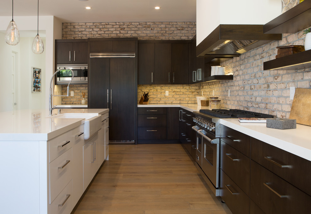 Photo of a modern l-shaped kitchen in Orange County with a farmhouse sink, flat-panel cabinets, dark wood cabinets, brick splashback, panelled appliances, light hardwood floors and with island.