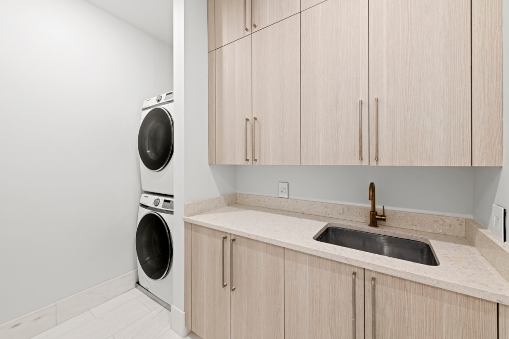Inspiration for a mid-sized modern single-wall dedicated laundry room in New York with an undermount sink, flat-panel cabinets, light wood cabinets, quartz benchtops, beige splashback, engineered quartz splashback, white walls, porcelain floors, a stacked washer and dryer, multi-coloured floor and beige benchtop.