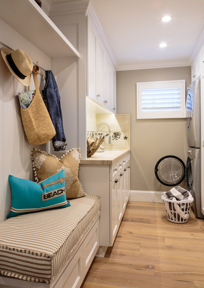 Photo of a large beach style galley dedicated laundry room in Orange County with a single-bowl sink, recessed-panel cabinets, white cabinets, limestone benchtops, grey walls, light hardwood floors and a stacked washer and dryer.