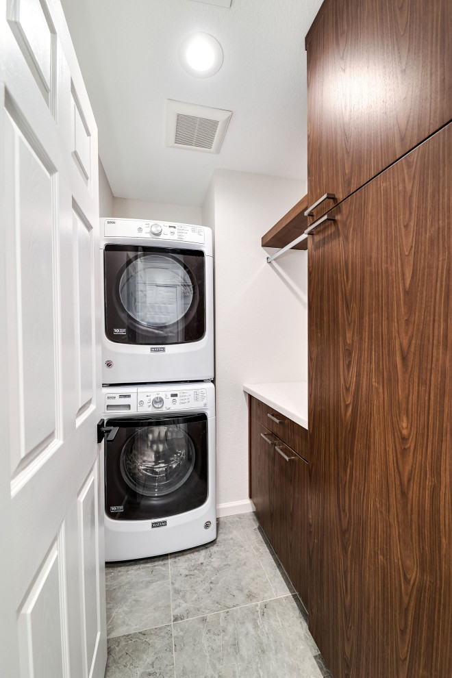 Photo of a small transitional u-shaped utility room in Phoenix with flat-panel cabinets, brown cabinets, quartz benchtops, white walls, porcelain floors, a stacked washer and dryer, grey floor and white benchtop.