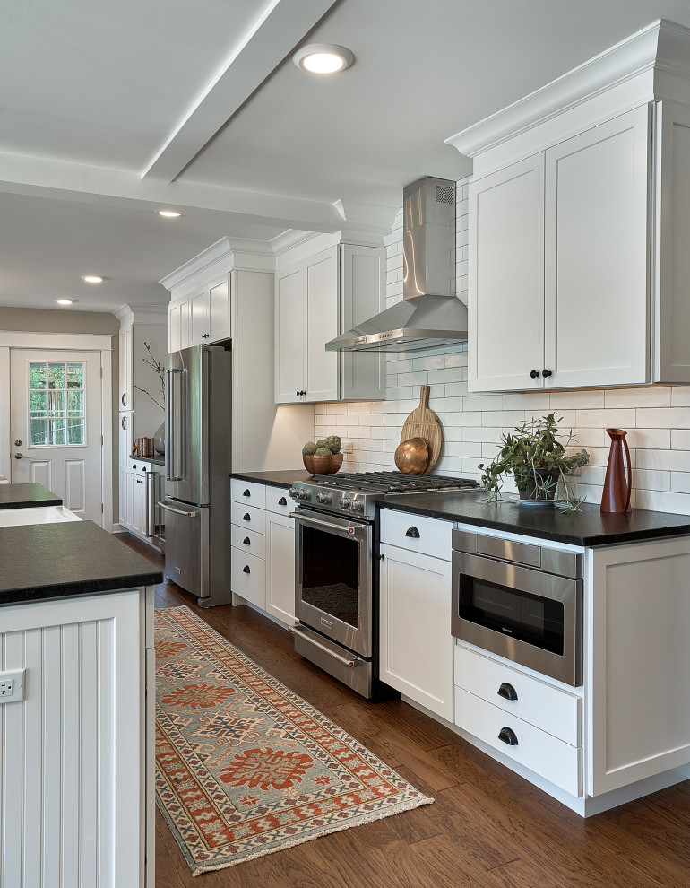 Photo of a mid-sized country galley eat-in kitchen in Chicago with a farmhouse sink, shaker cabinets, white cabinets, granite benchtops, white splashback, ceramic splashback, stainless steel appliances, medium hardwood floors, with island, brown floor, black benchtop and coffered.