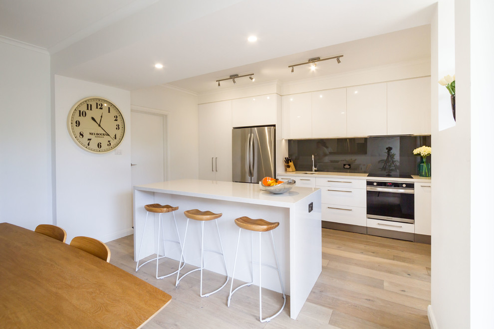 Photo of a mid-sized modern galley eat-in kitchen in Melbourne with flat-panel cabinets, white cabinets, quartz benchtops, grey splashback, glass sheet splashback, stainless steel appliances, light hardwood floors and with island.
