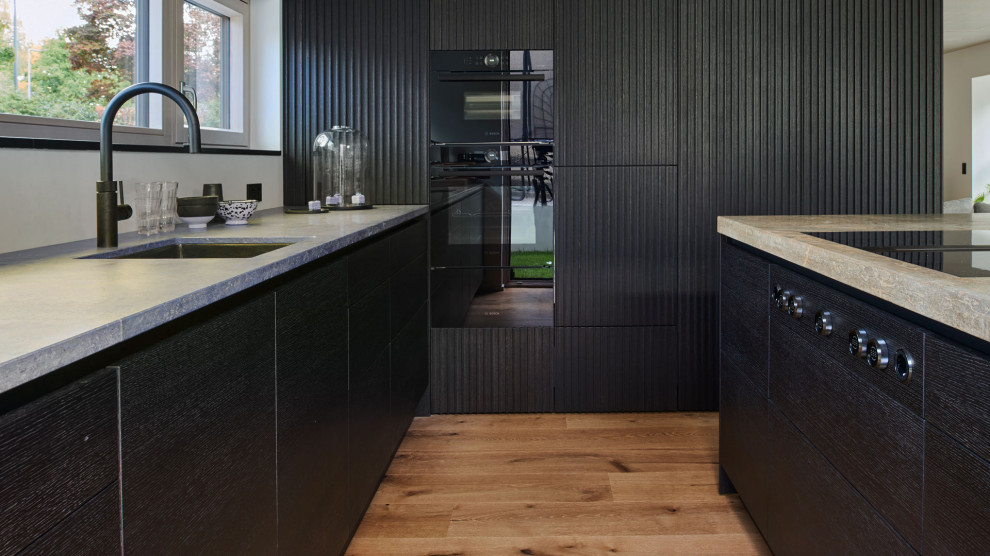 Moderne Küche mit Lamellenschränken, schwarzen Schränken, Granit-Arbeitsplatte, schwarzen Elektrogeräten, gebeiztem Holzboden, Kücheninsel, braunem Boden und beiger Arbeitsplatte in München