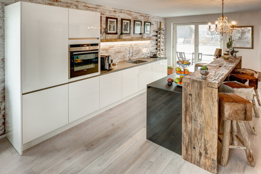 Inspiration for a large contemporary galley eat-in kitchen in Cologne with flat-panel cabinets, white cabinets, light hardwood floors, with island, an integrated sink, stainless steel benchtops, brown splashback and black appliances.