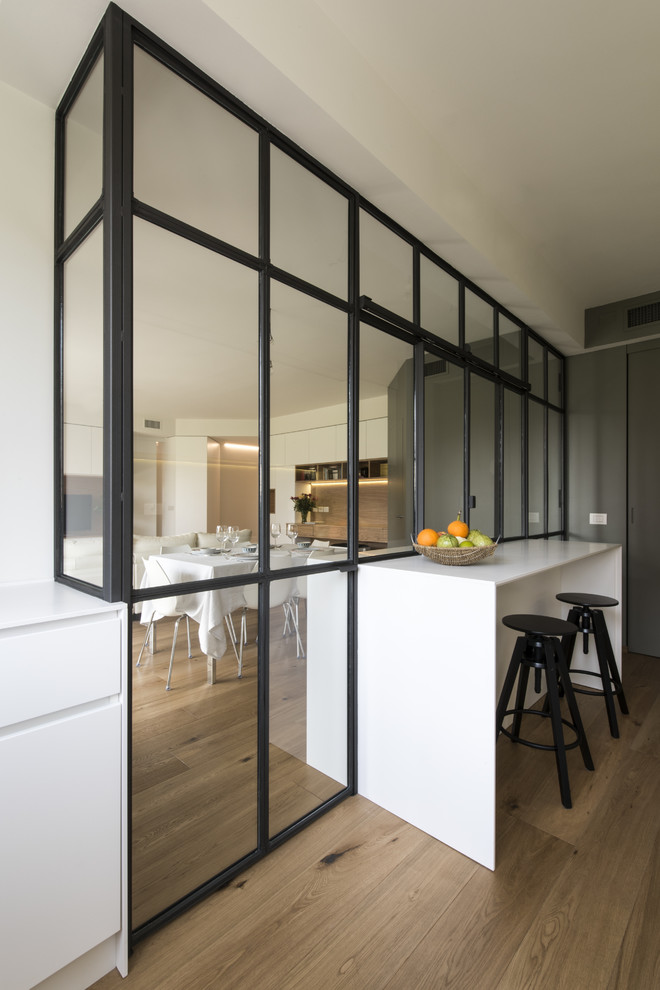 This is an example of a mid-sized modern galley separate kitchen in Milan with an undermount sink, flat-panel cabinets, white cabinets, solid surface benchtops, grey splashback, glass sheet splashback, stainless steel appliances, light hardwood floors, with island and beige floor.