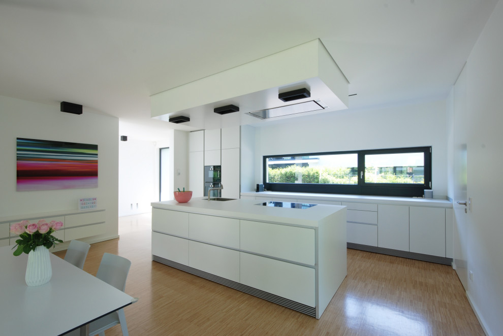 This is an example of a large modern eat-in kitchen in Dusseldorf with a single-bowl sink, flat-panel cabinets, white cabinets, laminate benchtops, white splashback, window splashback, black appliances, bamboo floors, with island, beige floor and white benchtop.