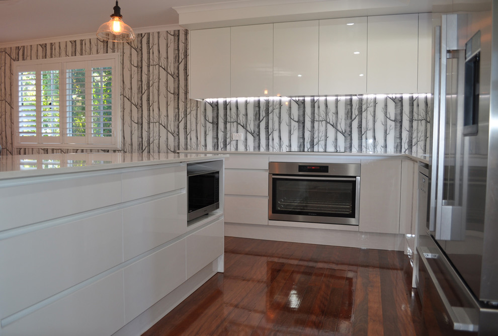 Photo of a mid-sized contemporary l-shaped eat-in kitchen in Brisbane with an undermount sink, flat-panel cabinets, white cabinets, quartz benchtops, multi-coloured splashback, glass sheet splashback, stainless steel appliances, dark hardwood floors and with island.