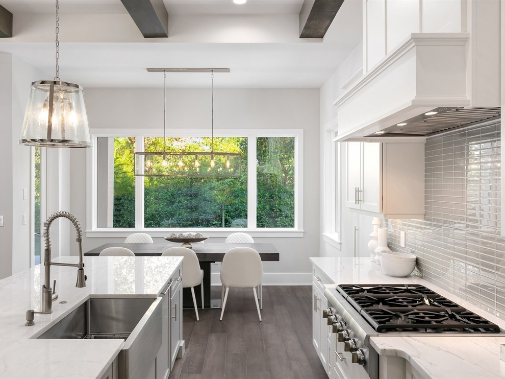 Large transitional l-shaped open plan kitchen in Orlando with a farmhouse sink, shaker cabinets, white cabinets, grey splashback, glass tile splashback, stainless steel appliances, dark hardwood floors, with island, brown floor, white benchtop and quartz benchtops.