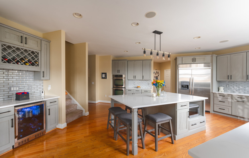Large transitional u-shaped medium tone wood floor and brown floor eat-in kitchen photo with a farmhouse sink, shaker cabinets, gray cabinets, quartz countertops, gray backsplash, glass tile backsplash, stainless steel appliances, an island and white countertops