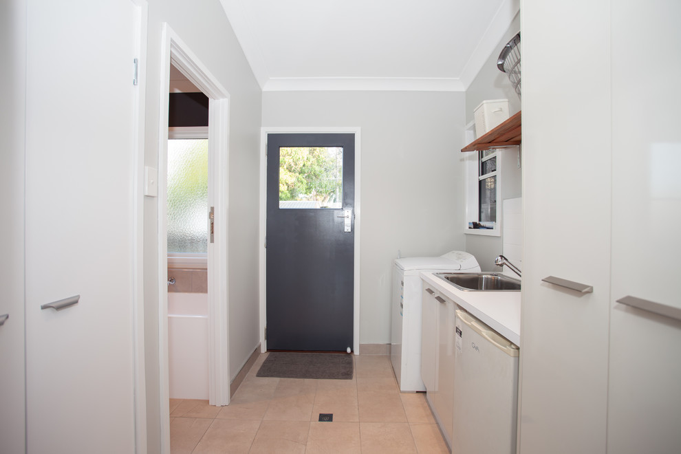 Photo of a small traditional single-wall dedicated laundry room in Other with a drop-in sink, laminate benchtops, grey walls, white cabinets, flat-panel cabinets, white splashback, subway tile splashback, ceramic floors, beige floor and white benchtop.