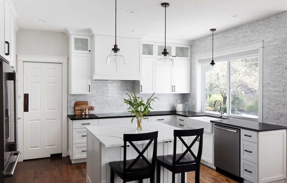 Mid-sized contemporary u-shaped eat-in kitchen in Portland with a farmhouse sink, shaker cabinets, white cabinets, granite benchtops, white splashback, marble splashback, stainless steel appliances, dark hardwood floors, with island, brown floor and black benchtop.