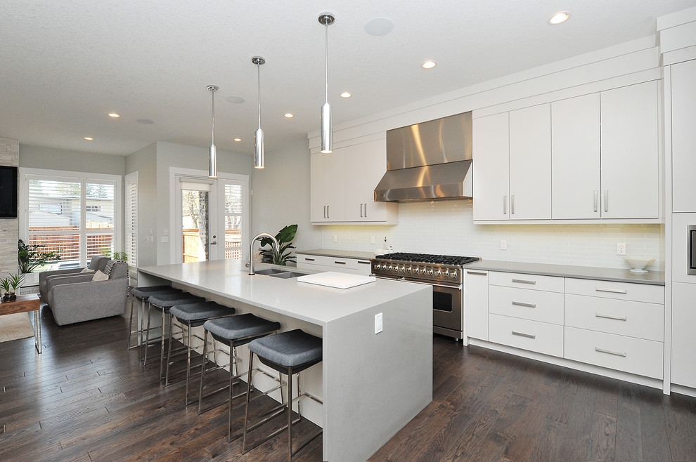 Large modern l-shaped open plan kitchen in Calgary with a double-bowl sink, flat-panel cabinets, white cabinets, concrete benchtops, white splashback, glass tile splashback, stainless steel appliances, dark hardwood floors, with island, brown floor and grey benchtop.