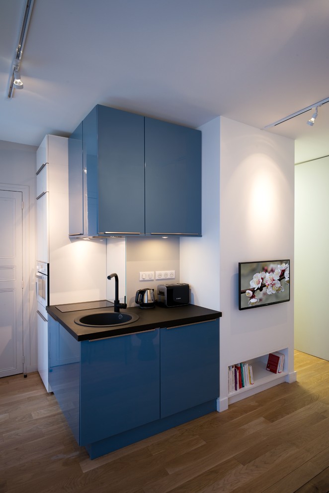 Photo of a small contemporary l-shaped open plan kitchen in Other with beaded inset cabinets, blue cabinets, no island, a drop-in sink, laminate benchtops, grey splashback, glass sheet splashback, black appliances and medium hardwood floors.