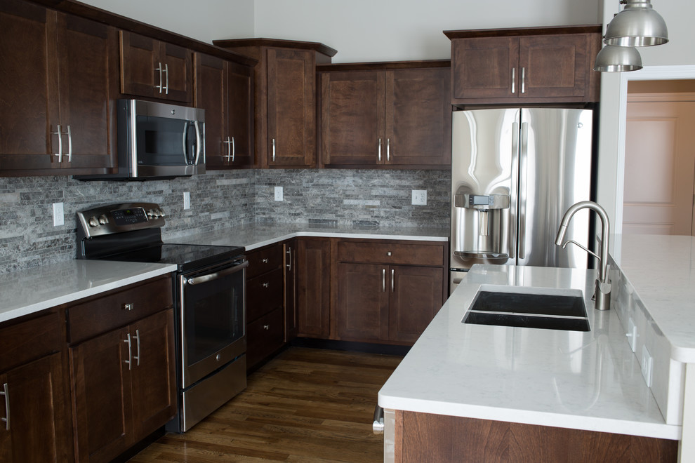 Mid-sized transitional l-shaped dark wood floor enclosed kitchen photo with a double-bowl sink, shaker cabinets, dark wood cabinets, quartz countertops, gray backsplash, stone tile backsplash, stainless steel appliances and an island