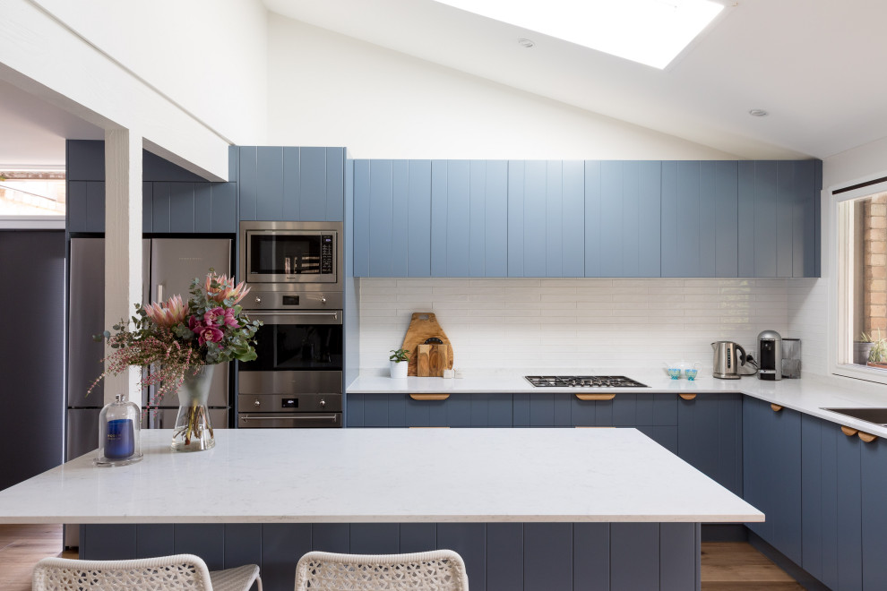 This is an example of a mid-sized midcentury kitchen with a double-bowl sink, blue cabinets, quartz benchtops, white splashback, porcelain splashback, stainless steel appliances, medium hardwood floors, brown floor and white benchtop.