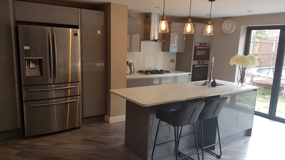 Medium sized contemporary single-wall open plan kitchen in West Midlands with an integrated sink, glass-front cabinets, grey cabinets, quartz worktops, white splashback, laminate floors, an island, grey floors and white worktops.