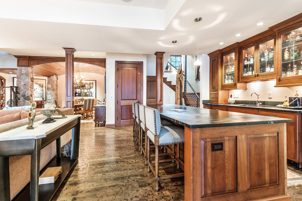 This is an example of a large country galley seated home bar in Milwaukee with medium wood cabinets, soapstone benchtops, an undermount sink, glass-front cabinets and dark hardwood floors.