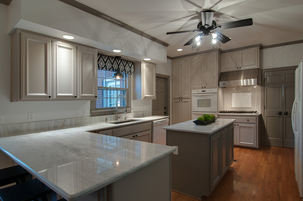 Photo of a mid-sized transitional l-shaped eat-in kitchen in Nashville with a farmhouse sink, recessed-panel cabinets, grey cabinets, granite benchtops, grey splashback, ceramic splashback, white appliances, medium hardwood floors and with island.