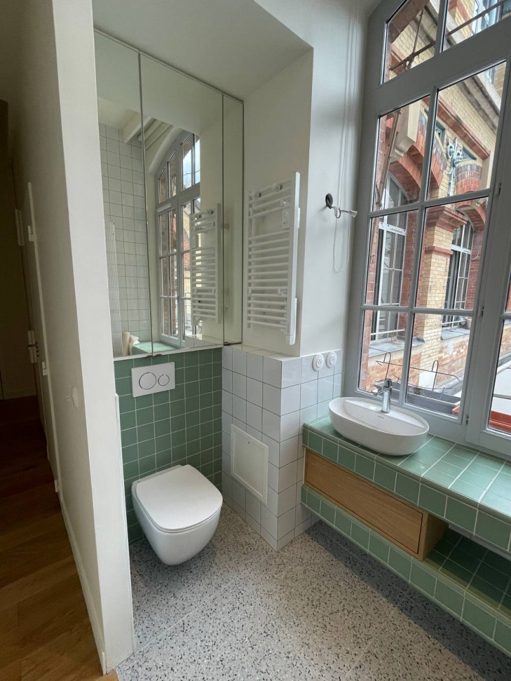 This is an example of a mid-sized contemporary 3/4 bathroom in Paris with beaded inset cabinets, a curbless shower, a wall-mount toilet, green tile, ceramic tile, green walls, terrazzo floors, a drop-in sink, tile benchtops, green floor, an open shower, green benchtops, a single vanity and a built-in vanity.