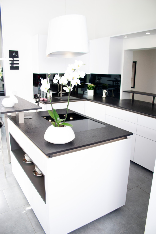 Offene, Kleine Moderne Küche in U-Form mit Waschbecken, flächenbündigen Schrankfronten, weißen Schränken, Küchenrückwand in Schwarz, Rückwand aus Spiegelfliesen, schwarzen Elektrogeräten, Keramikboden, Kücheninsel, grauem Boden und schwarzer Arbeitsplatte in München
