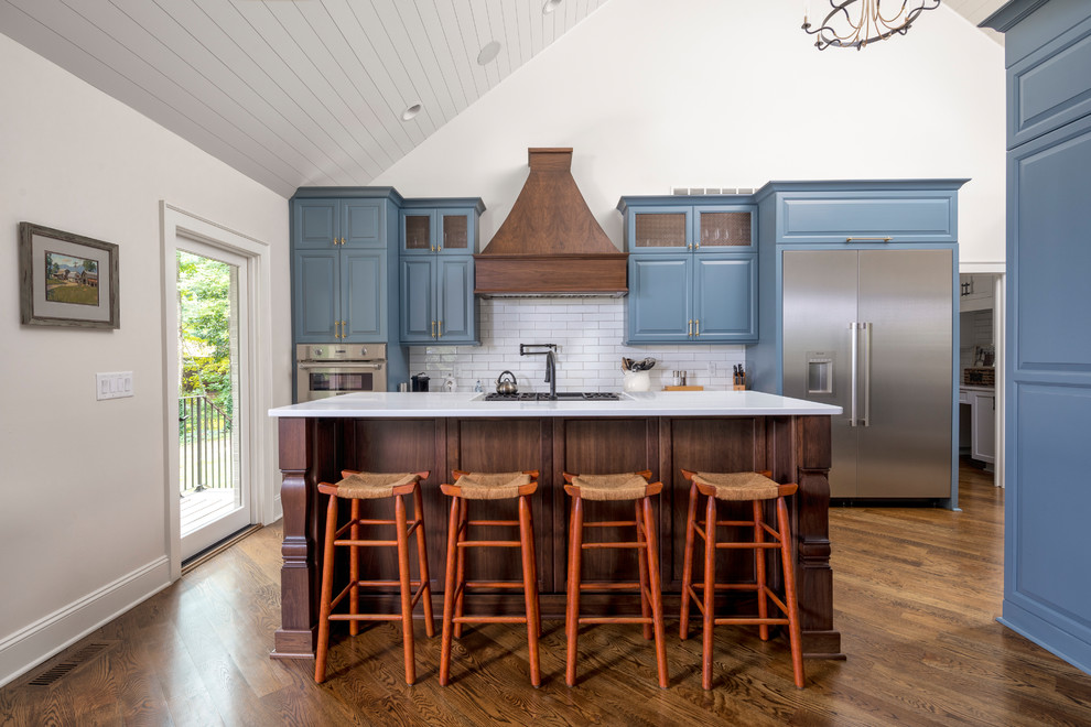Design ideas for a large transitional galley open plan kitchen in Charlotte with a farmhouse sink, blue cabinets, quartz benchtops, white splashback, subway tile splashback, stainless steel appliances, medium hardwood floors, with island, brown floor, white benchtop and raised-panel cabinets.