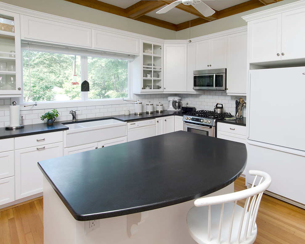 Inspiration for a mid-sized transitional u-shaped medium tone wood floor enclosed kitchen remodel with a farmhouse sink, shaker cabinets, white cabinets, granite countertops, white backsplash, ceramic backsplash, stainless steel appliances and an island