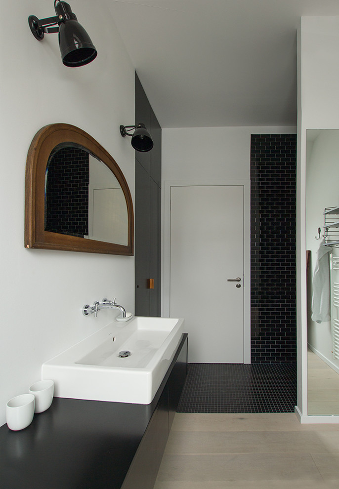 Photo of a mid-sized industrial 3/4 bathroom in Berlin with flat-panel cabinets, black cabinets, a curbless shower, a wall-mount toilet, black tile, porcelain tile, black walls, light hardwood floors, a vessel sink, solid surface benchtops, beige floor and an open shower.