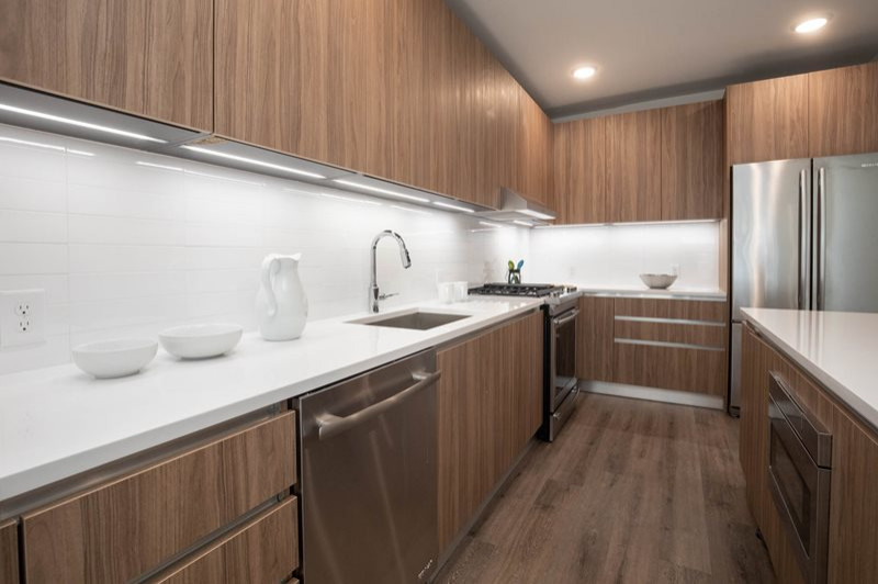 Bild på ett funkis vit vitt kök, med en undermonterad diskho, släta luckor, skåp i mellenmörkt trä, bänkskiva i kvarts, vitt stänkskydd, stänkskydd i porslinskakel, rostfria vitvaror, mellanmörkt trägolv och en köksö