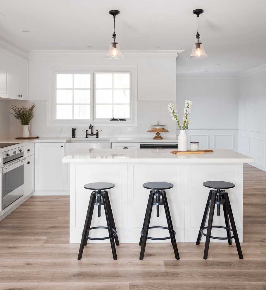 Inspiration for a mid-sized transitional l-shaped open plan kitchen in Sydney with a farmhouse sink, shaker cabinets, white cabinets, marble benchtops, white splashback, marble splashback, stainless steel appliances, laminate floors, with island, brown floor and white benchtop.