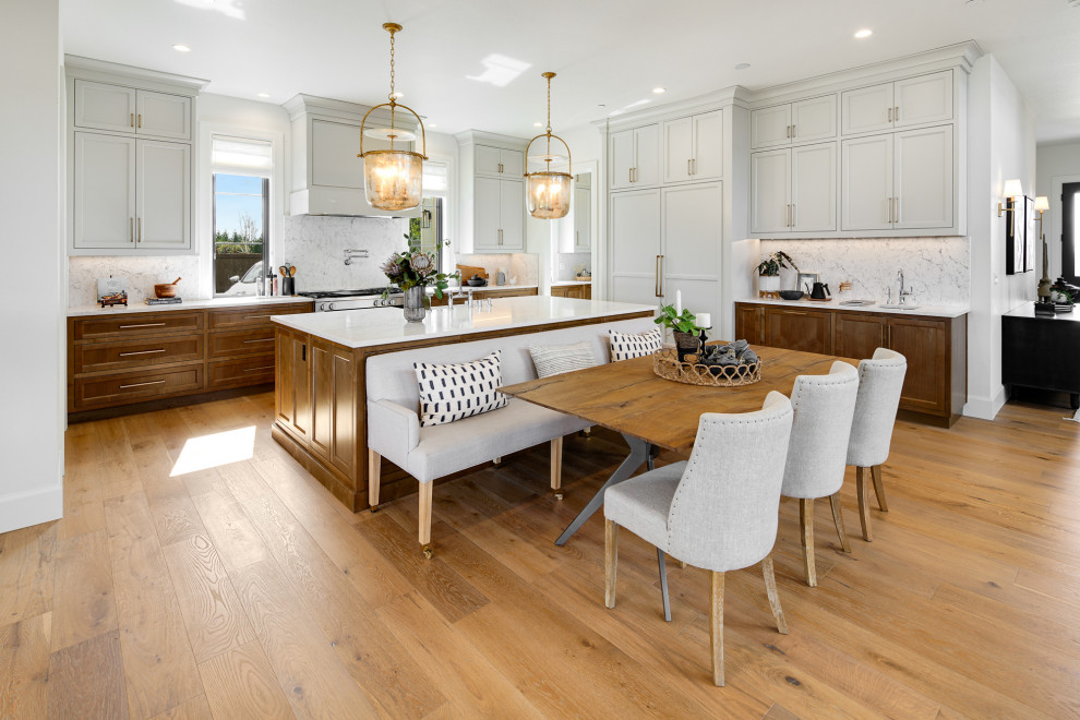 Transitional l-shaped open plan kitchen in Portland with a farmhouse sink, shaker cabinets, grey cabinets, white splashback, stone slab splashback, panelled appliances, medium hardwood floors, with island, brown floor and white benchtop.