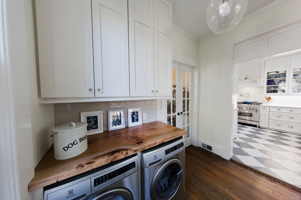 Mid-sized elegant medium tone wood floor and brown floor laundry room photo in Other with wood countertops, beige backsplash, white walls, a side-by-side washer/dryer and brown countertops
