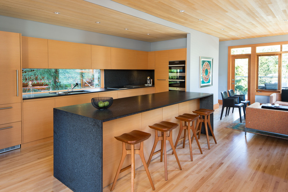 Inspiration for an expansive modern l-shaped open plan kitchen in Vancouver with flat-panel cabinets, granite benchtops, light hardwood floors, with island, window splashback, black appliances, beige floor, a drop-in sink, light wood cabinets, black splashback and black benchtop.