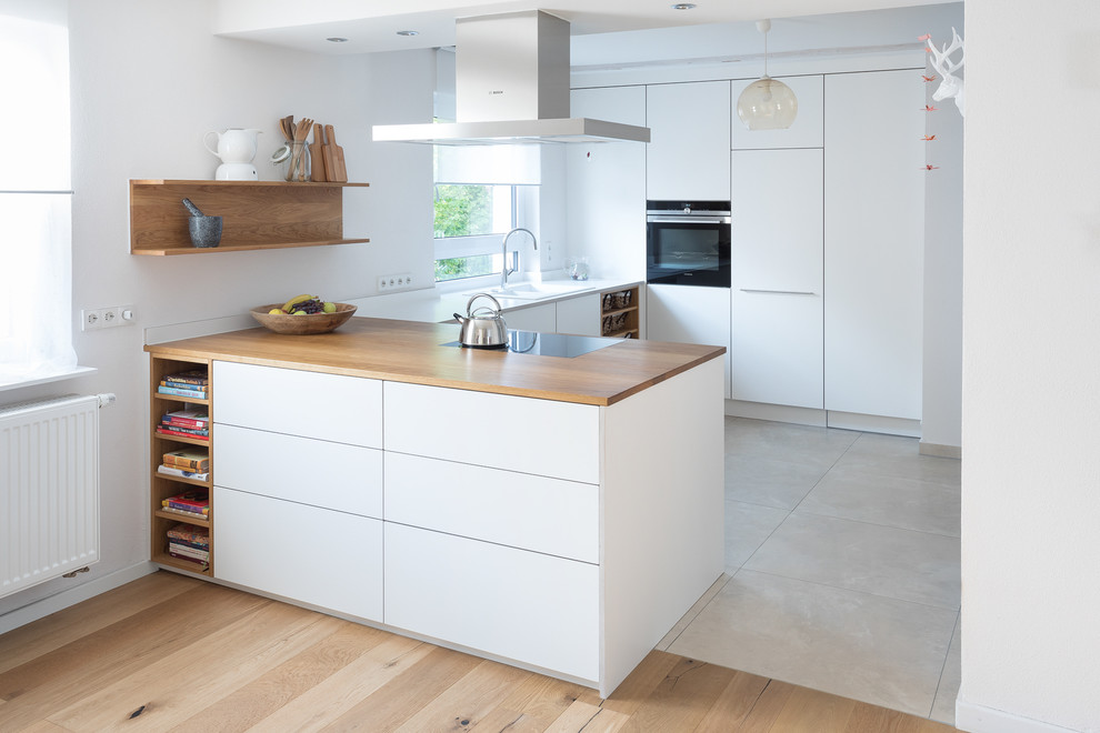 Inspiration for a small contemporary u-shaped open plan kitchen in Stuttgart with a drop-in sink, flat-panel cabinets, white cabinets, wood benchtops, white splashback, black appliances, limestone floors, beige floor, brown benchtop and a peninsula.