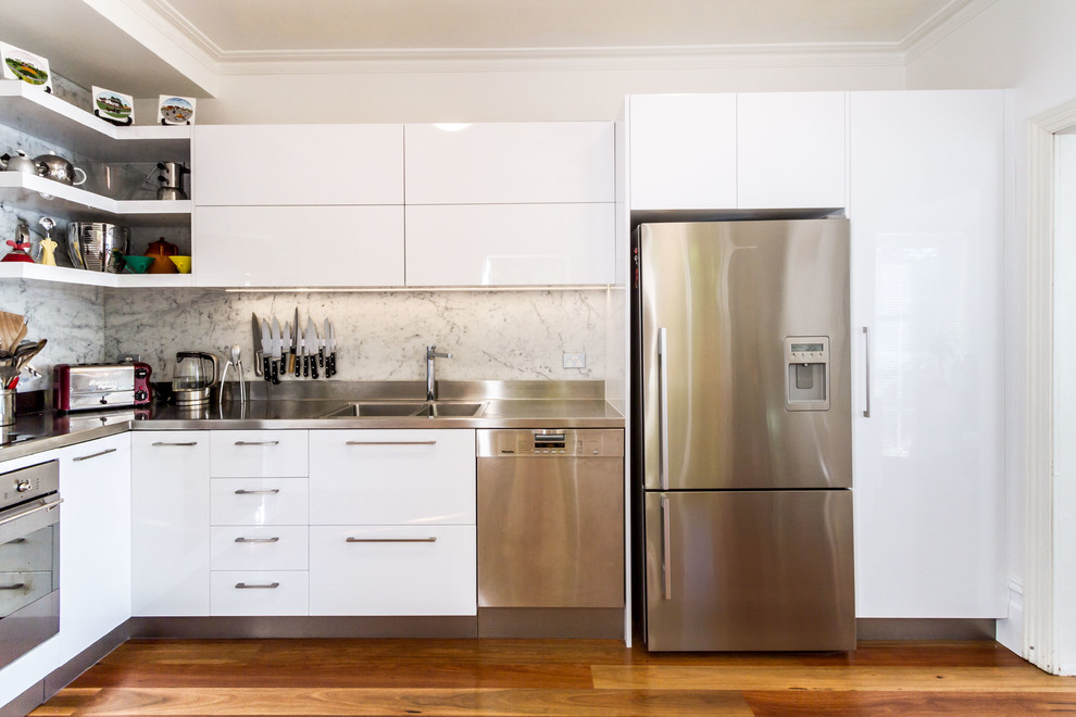 Small contemporary l-shaped kitchen pantry in Melbourne with an integrated sink, flat-panel cabinets, white cabinets, stainless steel benchtops, white splashback, marble splashback, stainless steel appliances, medium hardwood floors and brown floor.