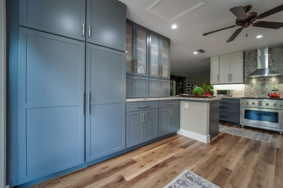 This is an example of a large country l-shaped kitchen pantry in Miami with a farmhouse sink, shaker cabinets, blue cabinets, granite benchtops, beige splashback, shiplap splashback, stainless steel appliances, vinyl floors, a peninsula, brown floor and multi-coloured benchtop.