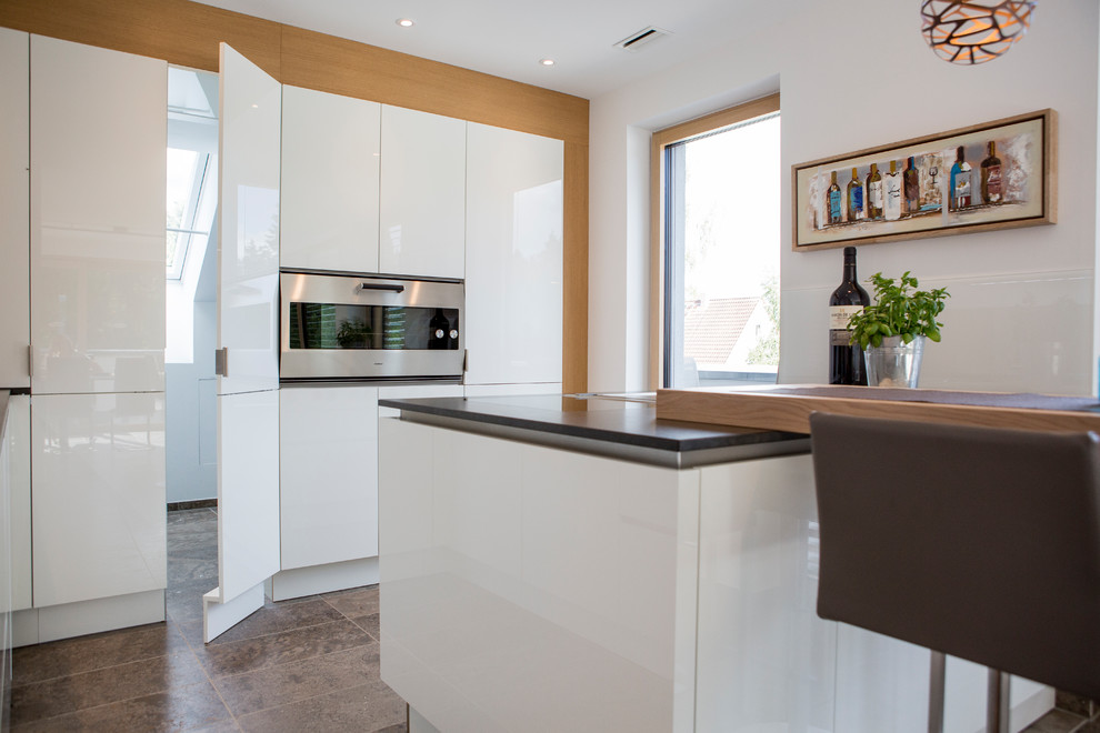 This is an example of a mid-sized contemporary l-shaped open plan kitchen in Frankfurt with a drop-in sink, flat-panel cabinets, white cabinets, granite benchtops, black splashback, glass sheet splashback, stainless steel appliances, terra-cotta floors, a peninsula, brown floor and black benchtop.
